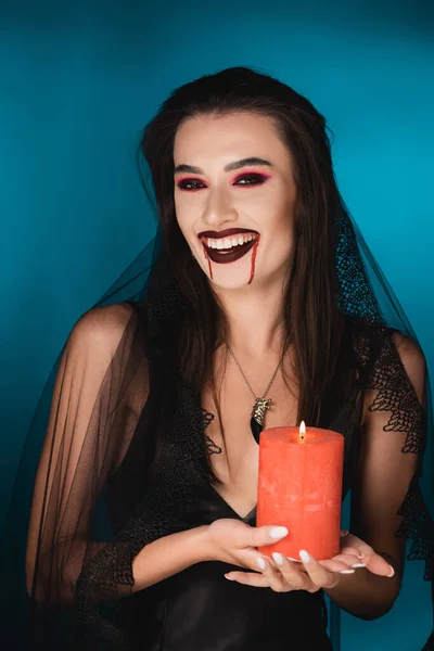 Woman in veil with blood on face holding red burning candle and laughing on blue — Stock Photo