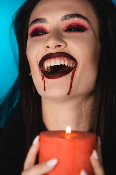 Foco seletivo de mulher morena com sangue no rosto segurando vela acesa e rindo no azul — Fotografia de Stock