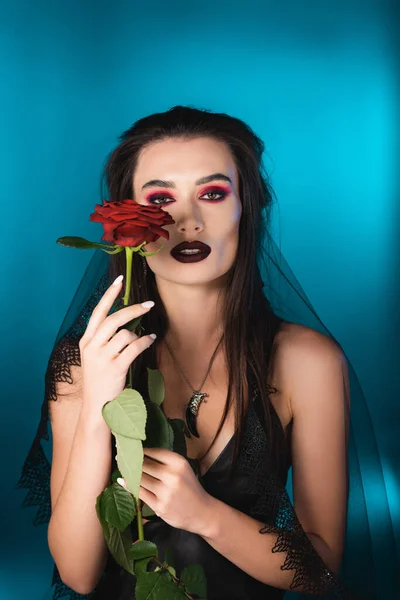 Morena mujer con maquillaje oscuro sosteniendo rosa roja en azul - foto de stock