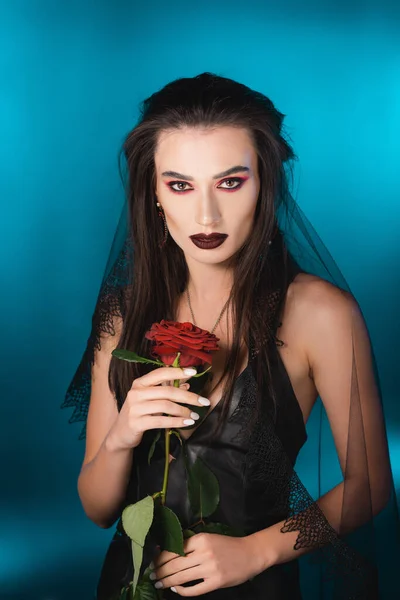 Joven morena con maquillaje oscuro sosteniendo rosa roja sobre azul - foto de stock