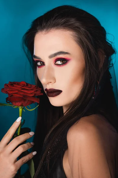 Jeune femme brune avec maquillage sombre regardant la caméra près de rose rouge sur bleu — Photo de stock