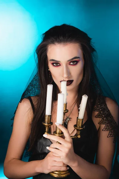 Femme pâle avec maquillage noir tenant des bougies allumées sur bleu — Photo de stock
