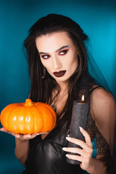 Joven mujer pálida con maquillaje negro y velo sosteniendo vela ardiente y calabaza en azul - foto de stock