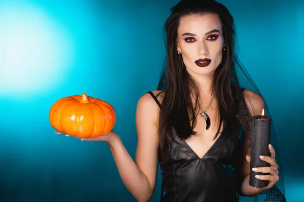 Young pale woman with black makeup and veil holding  candle and pumpkin on blue — Stock Photo