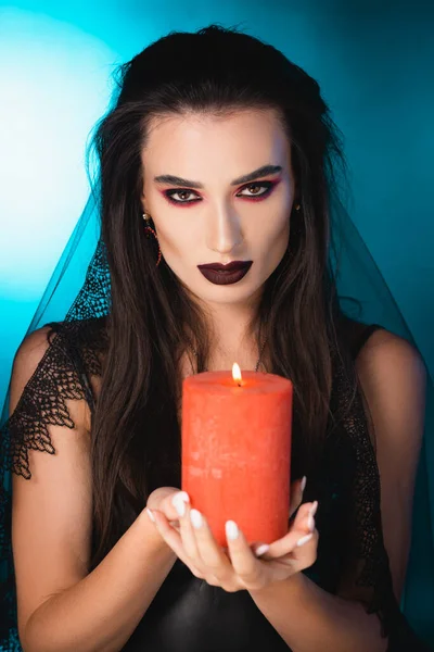 Pale woman with black makeup and veil holding burning candle on blue — Stock Photo