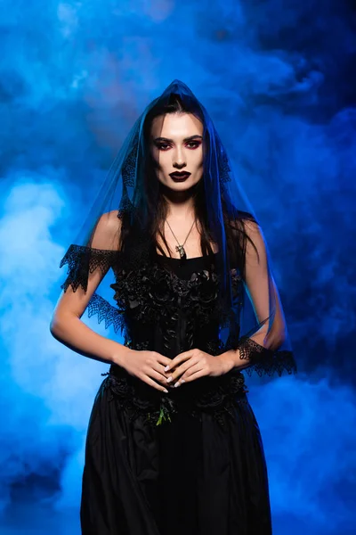 Bride in black dress and veil on blue with smoke, halloween concept — Stock Photo