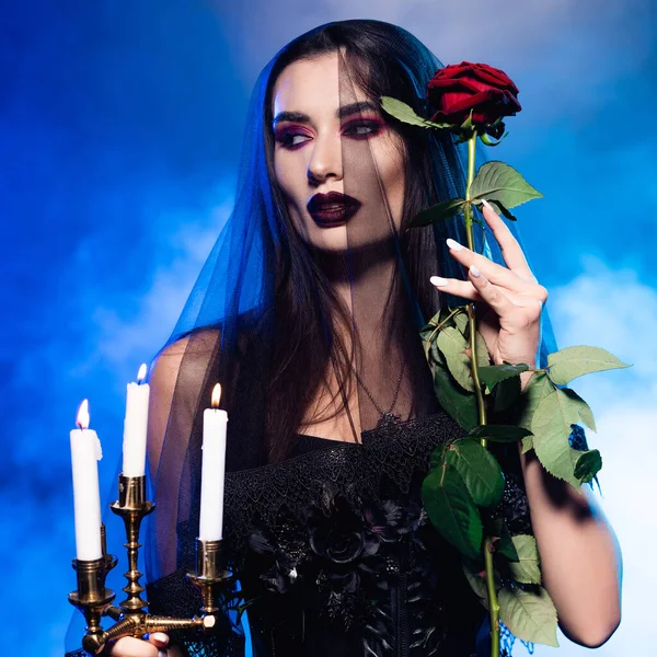 Woman in black dress and veil holding red rose and burning candles on blue with smoke, halloween concept — Stock Photo