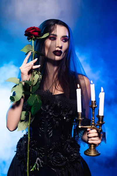Young woman in black dress and veil holding rose and burning candles on blue with smoke, halloween concept — Stock Photo