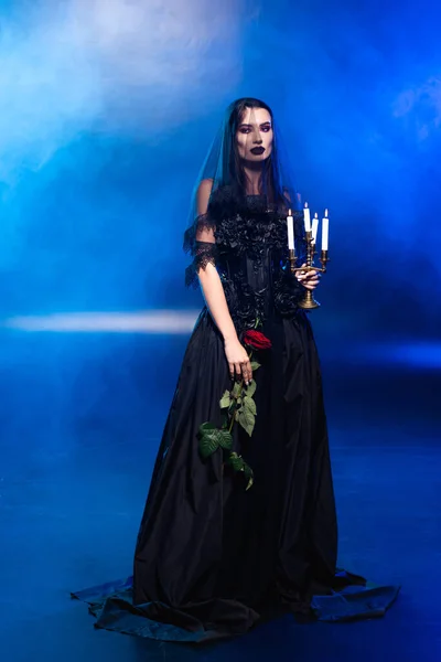 Bride in black dress and veil holding rose and candles on blue with smoke, halloween concept — Stock Photo