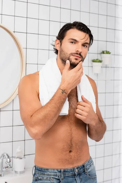 Hemdloser Mann berührt Handtuch und Kinn, während er im Badezimmer in die Kamera schaut — Stockfoto