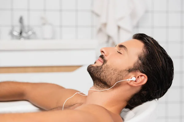 Hombre barbudo escuchando música en los auriculares en el baño - foto de stock