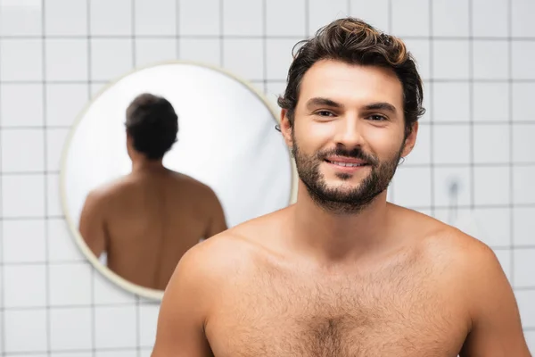 Hombre sin camisa sonriendo a la cámara con espejo sobre fondo borroso en el baño - foto de stock