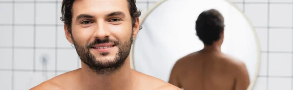 Бородатий і без сорочки чоловік посміхається на камеру біля дзеркала на розмитому фоні у ванній кімнаті, банер — стокове фото