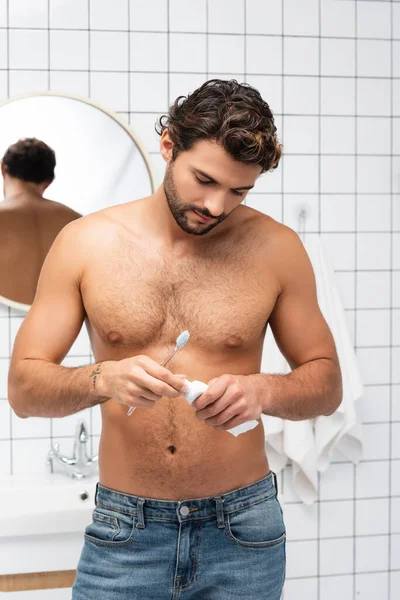 Мужчина без рубашки в джинсах держит зубную пасту и зубную щетку в ванной комнате — стоковое фото