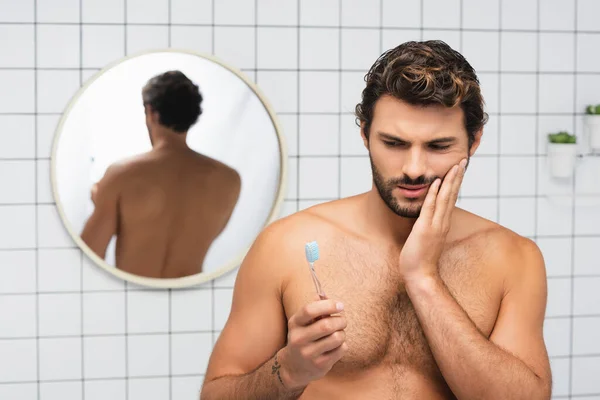 Uomo senza maglietta che tiene lo spazzolino da denti mentre tocca la guancia dolorosa in bagno — Foto stock