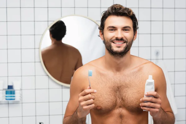 Веселий без сорочки чоловік дивиться на камеру, тримаючи зубну пасту і зубну щітку у ванній — стокове фото