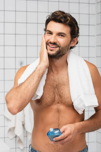 Hombre musculoso con toalla alrededor del cuello sonriendo mientras se aplica después de afeitarse loción en el baño - foto de stock