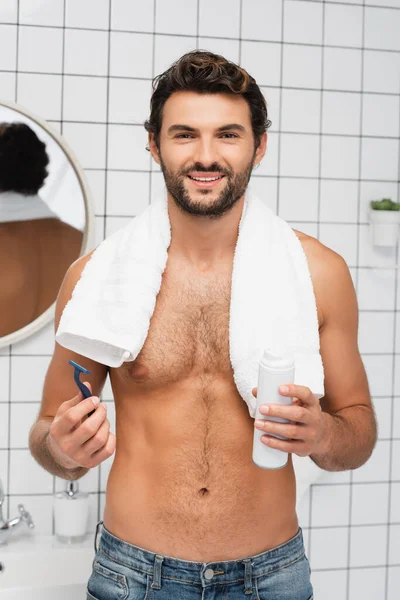 Hombre sonriente con toalla alrededor del cuello sosteniendo la afeitadora y espuma de afeitar en el baño - foto de stock