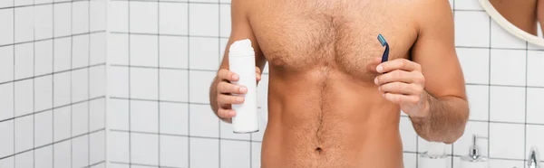 Vista recortada del hombre musculoso sosteniendo espuma de afeitar y afeitadora en el baño, pancarta - foto de stock