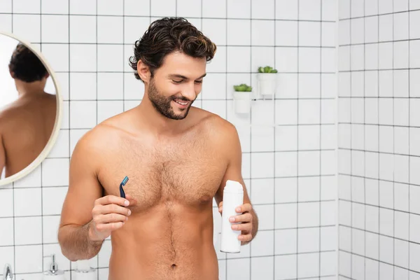 Hombre sin camisa sonriendo mientras sostiene espuma de afeitar y afeitadora desechable en el baño - foto de stock