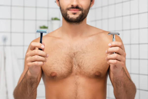 Vista cortada de lâminas de barbear em mãos de homem barbudo no fundo borrado no banheiro — Fotografia de Stock