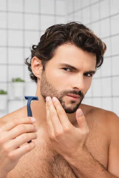 Бородатый мужчина держит бритву и смотрит в камеру в ванной комнате — стоковое фото