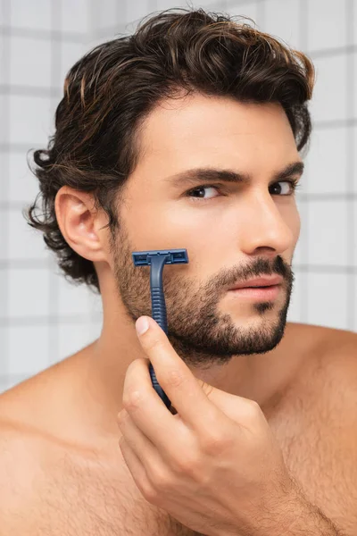 Hombre sin camisa mirando a la cámara mientras se afeita en el baño - foto de stock