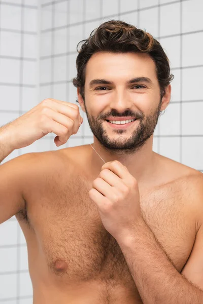 Hombre sin camisa sonriendo a la cámara mientras sostiene el hilo dental en el baño - foto de stock
