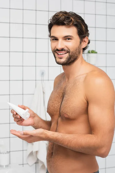 Sonriente hombre sin camisa sosteniendo tubo con crema cosmética en el baño - foto de stock