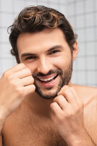 Sonriente hombre sin camisa usando hilo dental mientras mira a la cámara en el baño - foto de stock