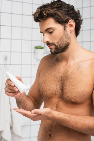 Joven hombre sin camisa sosteniendo tubo con crema cosmética en el baño - foto de stock