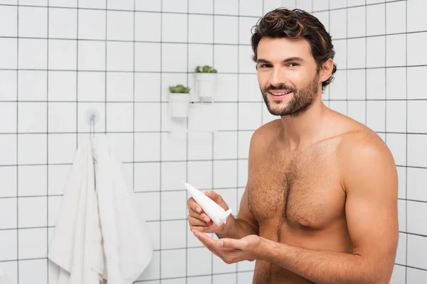 Hombre sin camisa sonriendo a la cámara mientras sostiene el tubo con crema cosmética en el baño - foto de stock