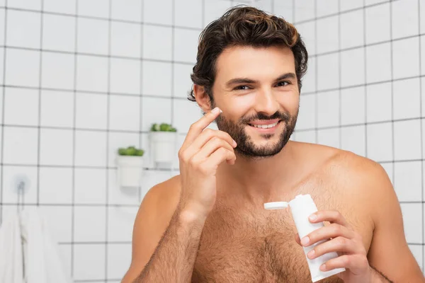 Uomo senza maglietta sorridente mentre applica la crema cosmetica in bagno — Foto stock