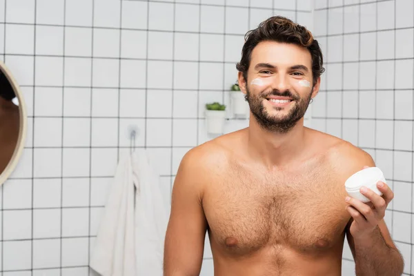 Sorrindo sem camisa homem segurando frasco com creme cosmético no banheiro — Fotografia de Stock