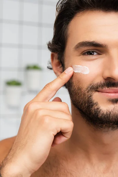 Vista recortada del hombre sonriente aplicando crema cosmética en la mejilla - foto de stock