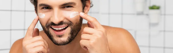 Hemdloser Mann lächelt in die Kamera, während er kosmetische Creme auf das Gesicht aufträgt, Banner — Stockfoto