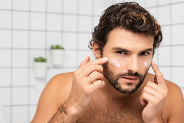 Бородатый мужчина наносит косметический крем на лицо в ванной комнате — стоковое фото
