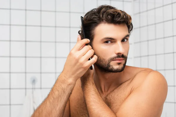 Hombre sin camisa mirando a la cámara mientras peina el pelo en el baño - foto de stock