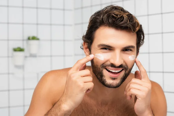 Homem alegre sem camisa aplicando creme facial e olhando para a câmera no banheiro — Fotografia de Stock