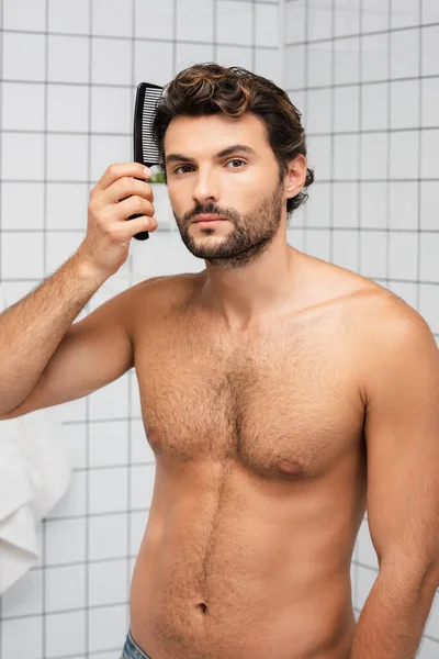 Homem muscular pentear o cabelo e olhando para a câmera no banheiro — Fotografia de Stock