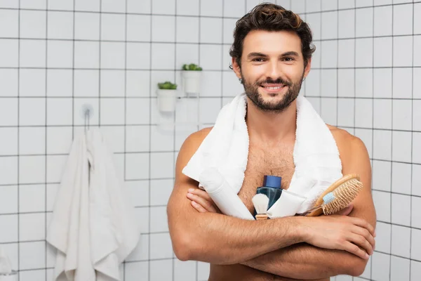 Hombre sonriente con toalla alrededor del cuello sosteniendo peine, pasta de dientes con cepillo de dientes y después de afeitarse loción en el baño - foto de stock