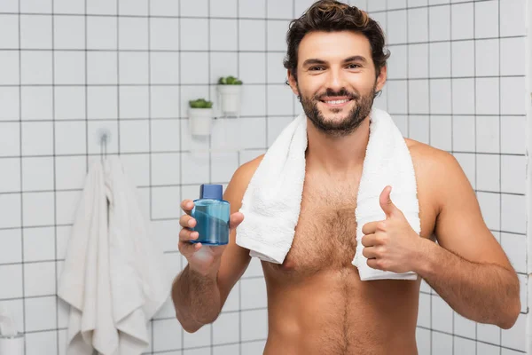 Smiling man with towel around neck showing thumb up and holding after shaving lotion in bathroom — Stock Photo