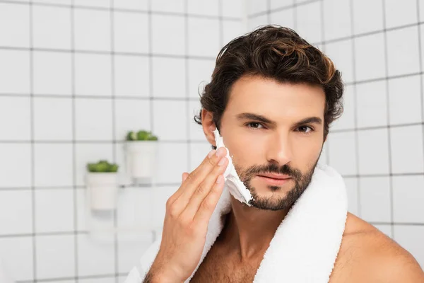 Hombre barbudo con toalla aplicando espuma de afeitar en el baño - foto de stock