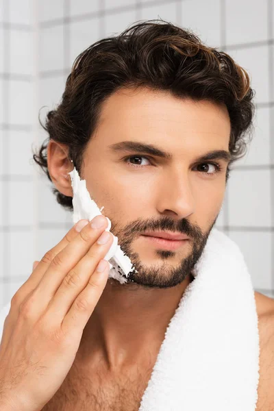 Homem sem camisa com toalha aplicando espuma de barbear e olhando para a câmera — Fotografia de Stock