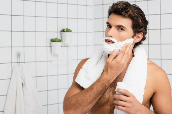 Мускулистый человек с полотенцем вокруг шеи наносит пену для бритья в ванной комнате — стоковое фото