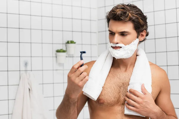 Hombre con espuma de afeitar en la cara que sostiene la cuchilla y la toalla en el baño - foto de stock