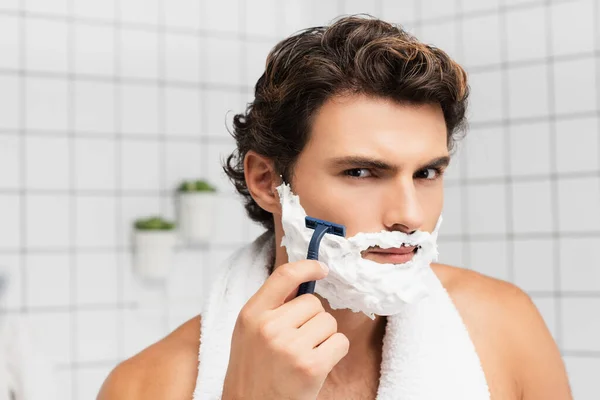 Joven con espuma de afeitar en la cara que sostiene la afeitadora desechable en el baño - foto de stock