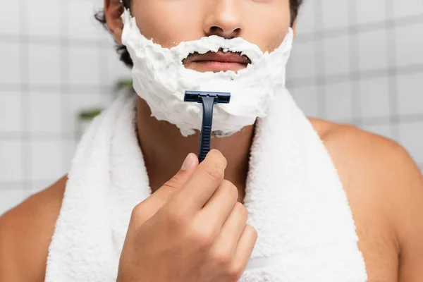 Cropped view of young man with shaving foam on face and towel around neck holding razor — Stock Photo