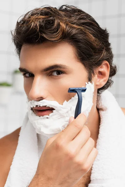 Brunette man with shaving foam on face shaving with razor in bathroom — Stock Photo