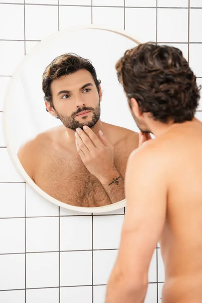 Чоловік без сорочки торкається підборіддя, дивлячись на дзеркало у ванній — стокове фото